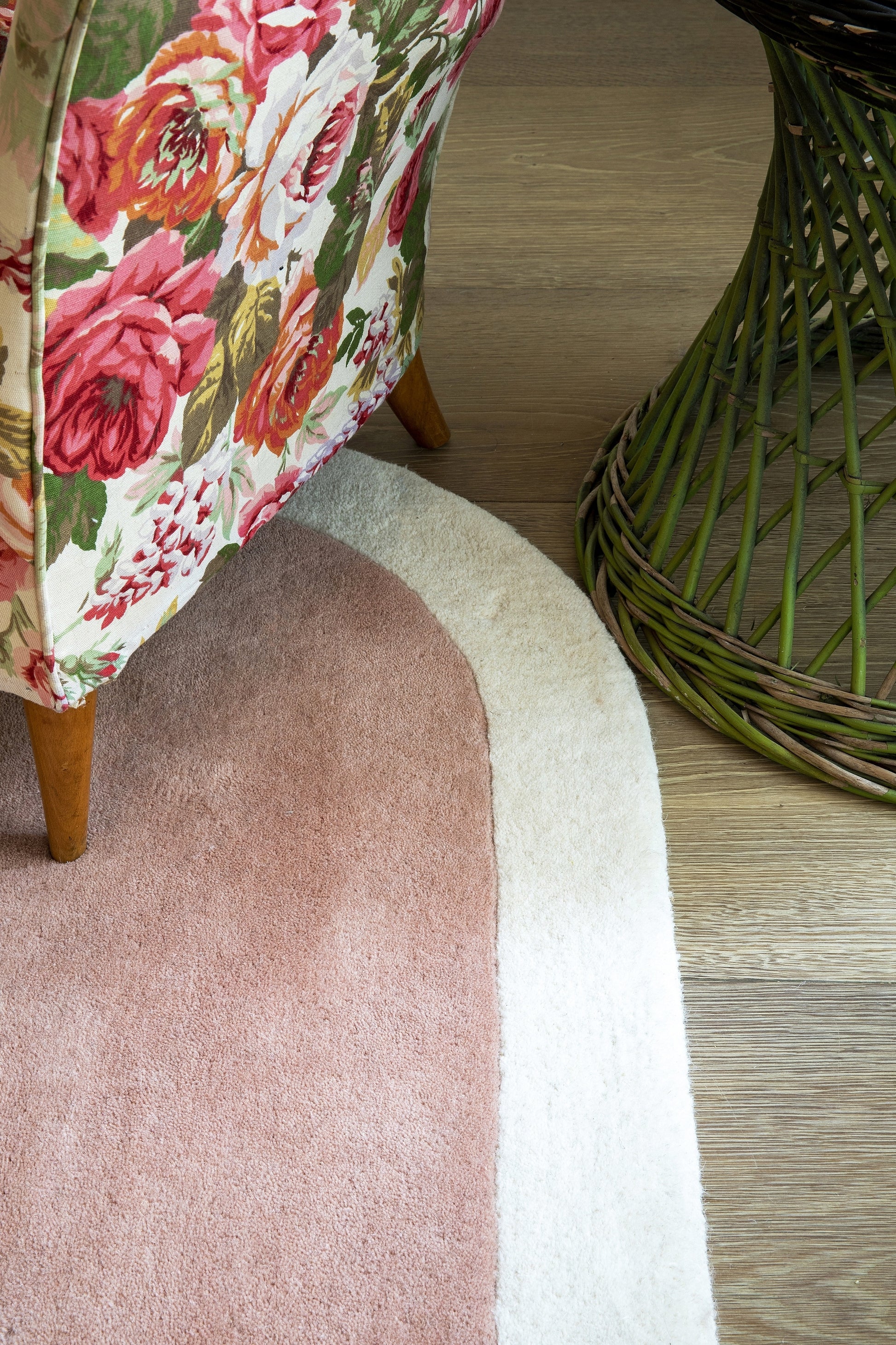 Floral pink & red upholstered chair with wooden legs beside a pink circular rug on a wood floor and a green wicker piece.