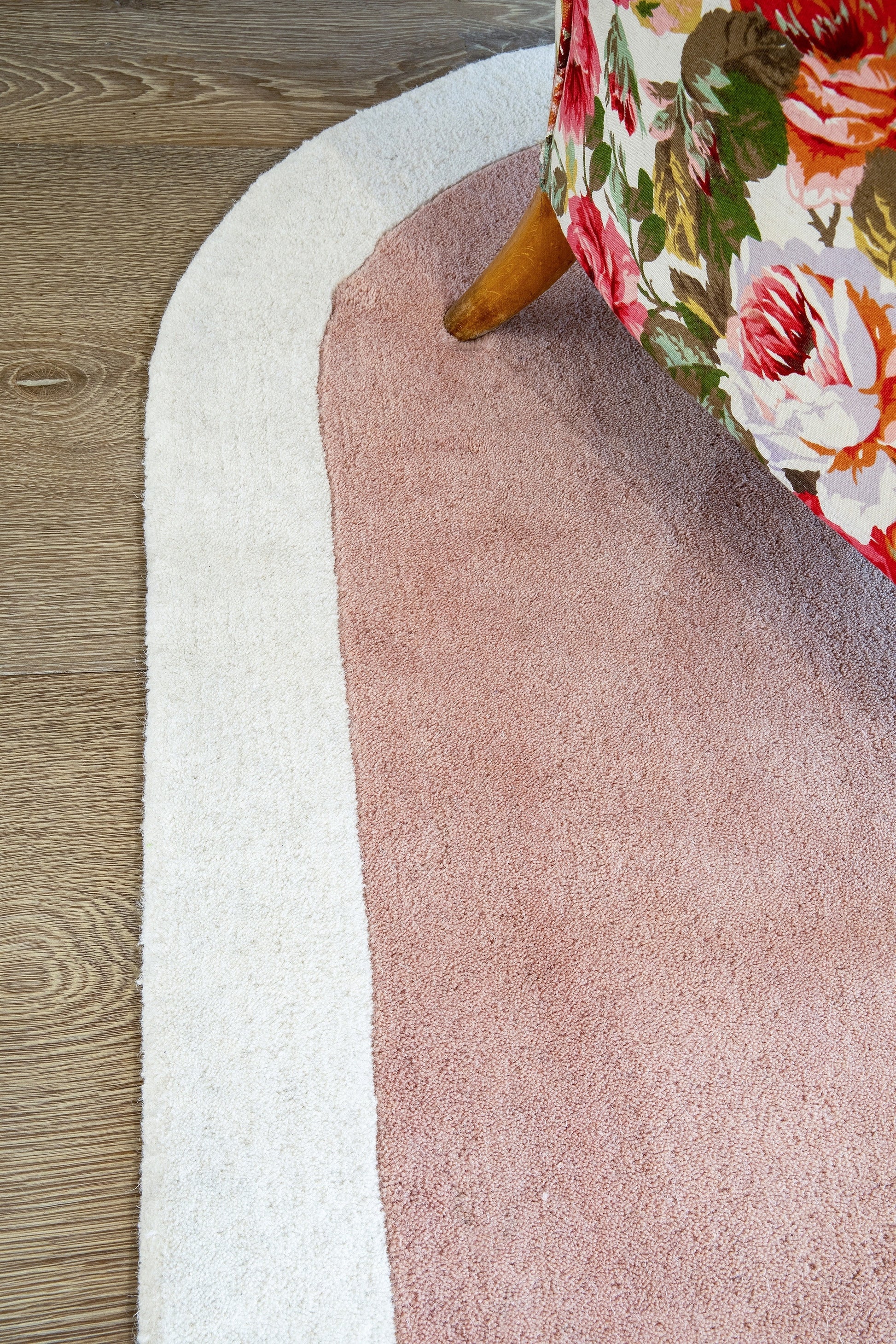 Oval pink center rug with white border on wood floor; floral upholstered chair leg peeks in.