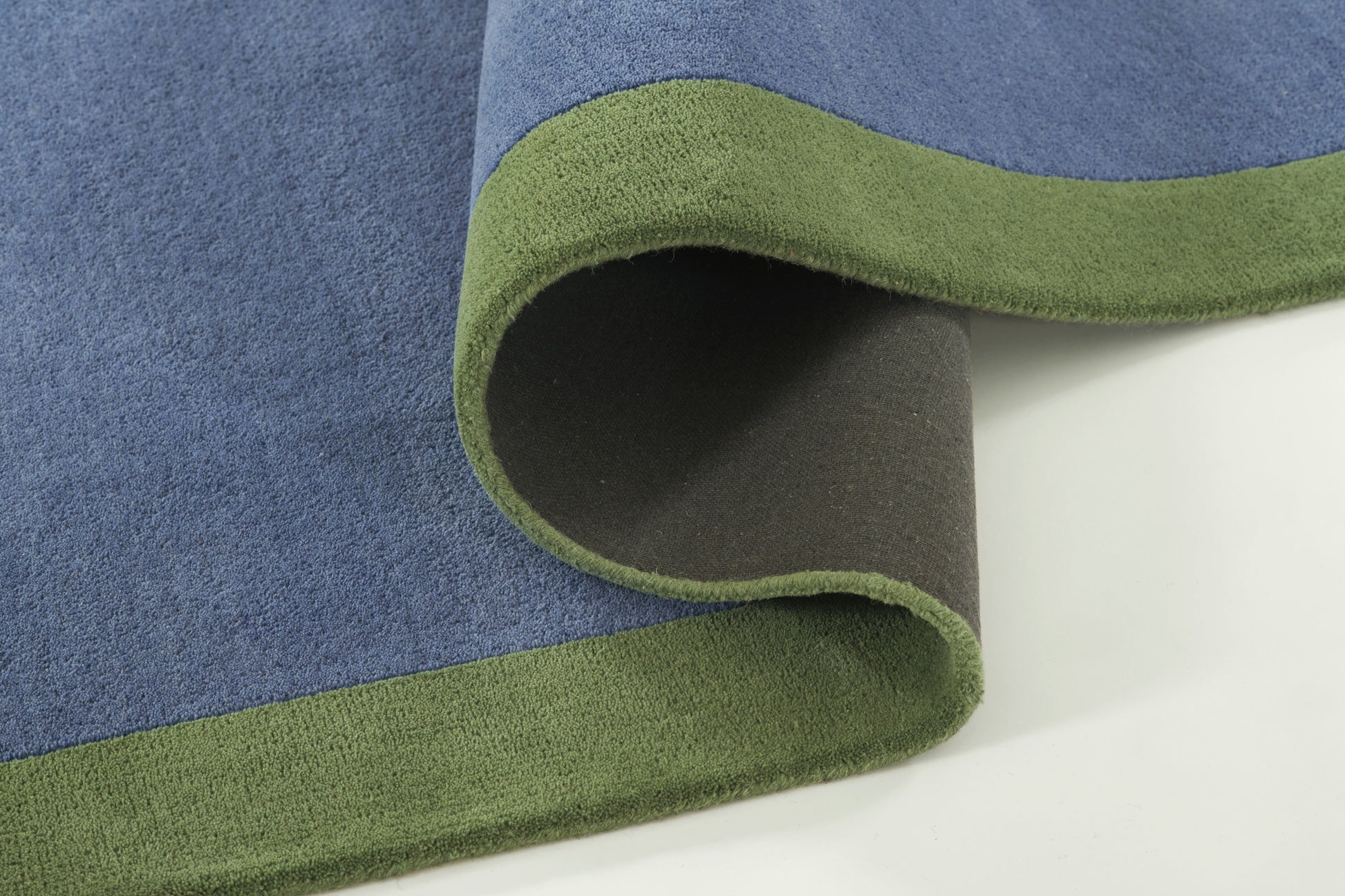 Blue rug with olive-green border, plush pile; rolled corner reveals dark backing.