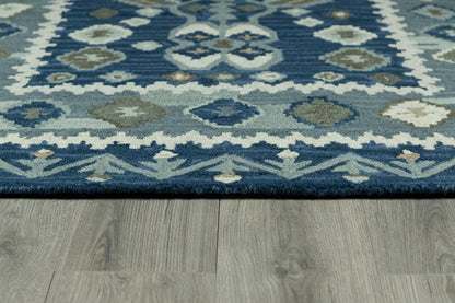 Blue geometric area rug with ivory and teal diamond motifs and navy border on gray wood floor.