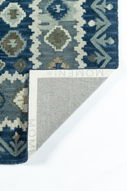 Rectangular blue-teal geometric rug with diamond motifs and beige border; folded corner reveals grey woven back and Momeni edge.