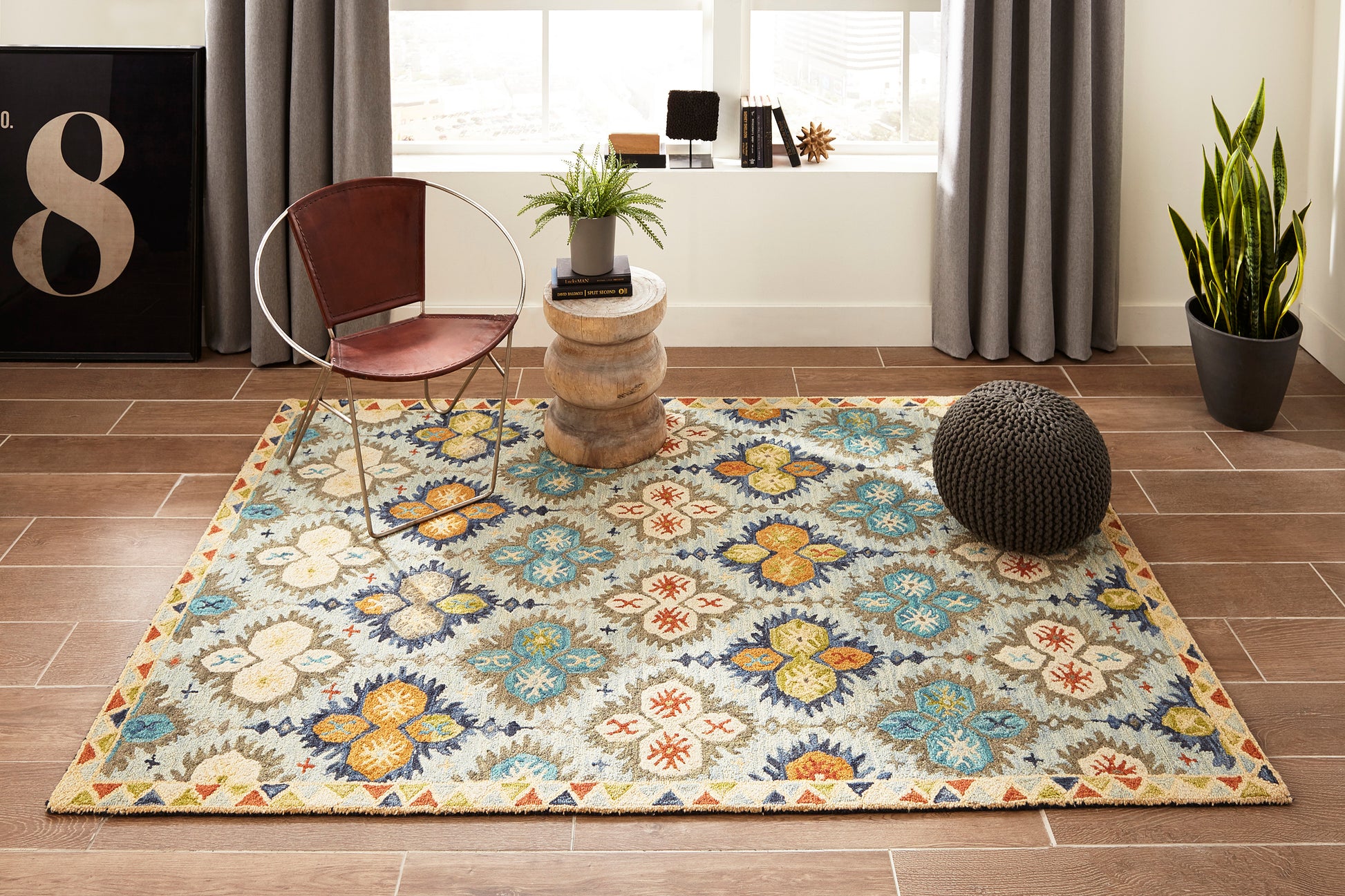 Colorful floral patterned rug on wood floor with brown leather chair, wooden side table, knitted pouf, snake plant.