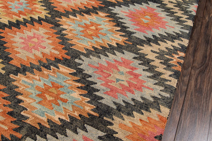 Geometric kilim rug with loud orange, teal, peach, red zigzag diamonds on dark background, edge of wood floor.