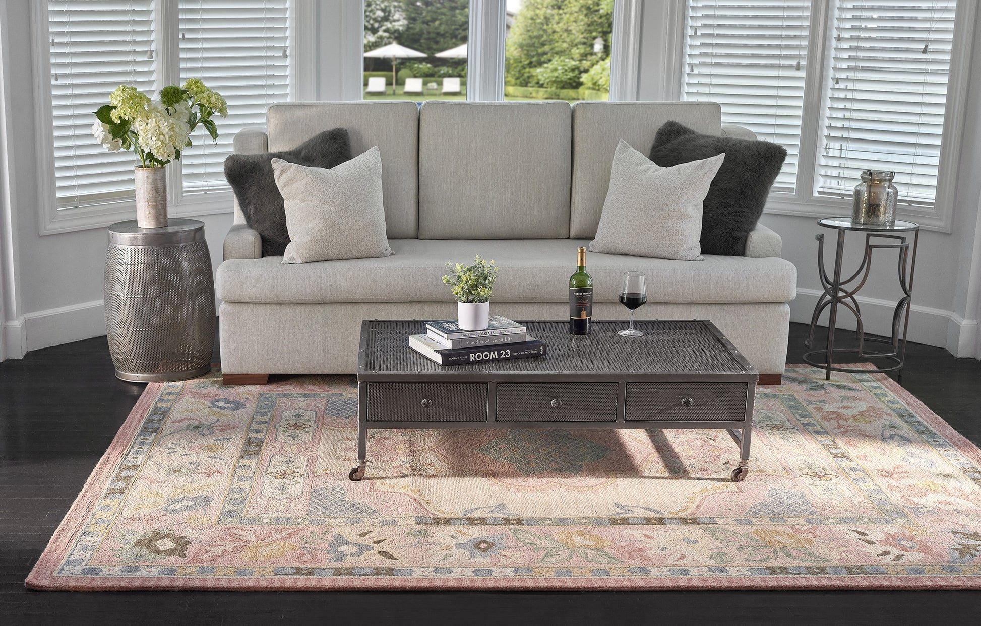 Pastel floral rug (8' x 11') in light gray tones in a living room, paired with a light gray sofa, dark metal coffee table, wine bottle & glass, and a plant.