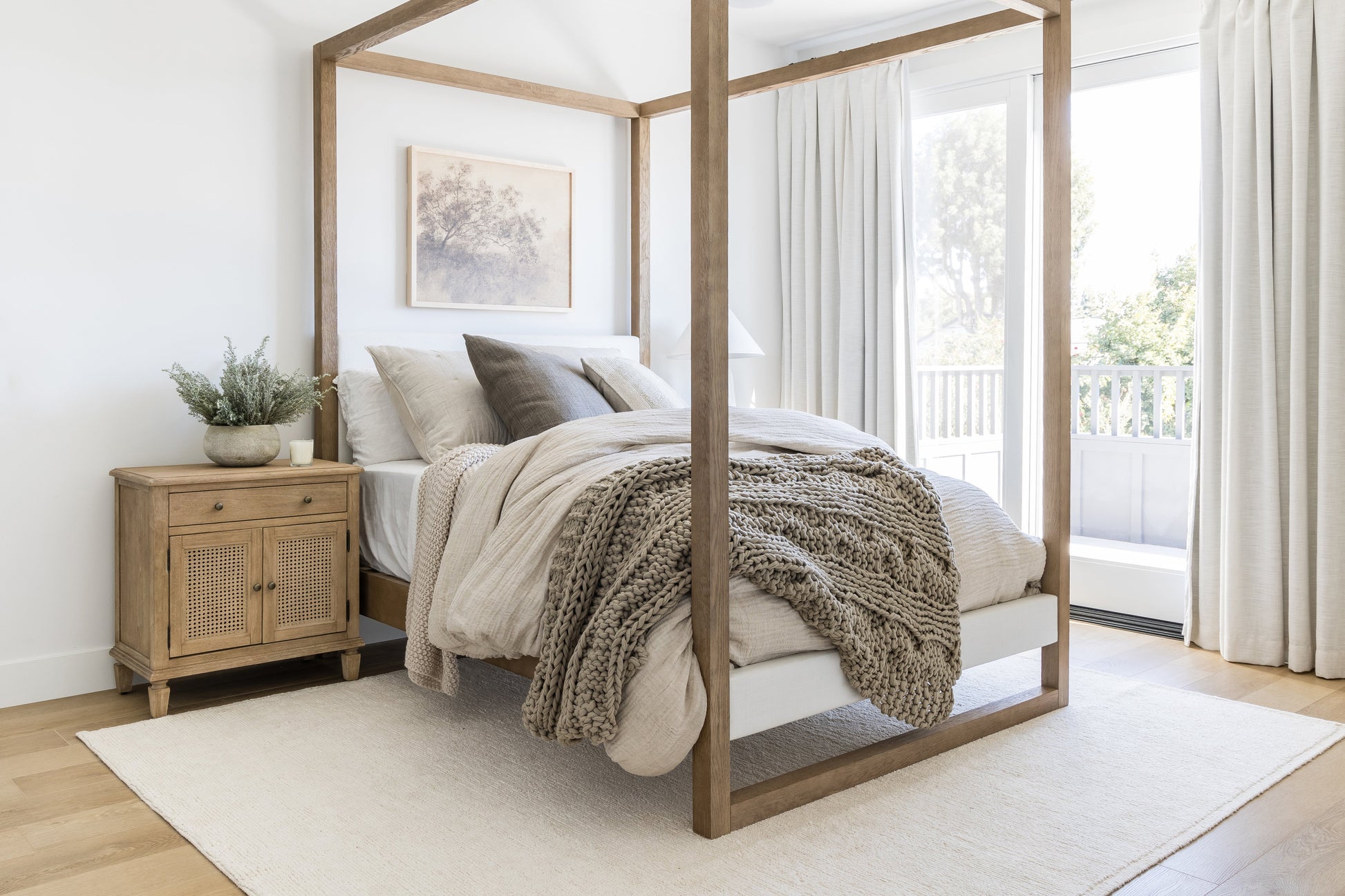 Neutral bedroom with a wooden four-poster bed, chunky knit throw, beige linens, wicker nightstand, plant, art, and windows.