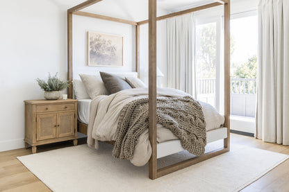 Neutral bedroom with a wooden four-poster bed, chunky knit throw, beige linens, wicker nightstand, plant, art, and windows.