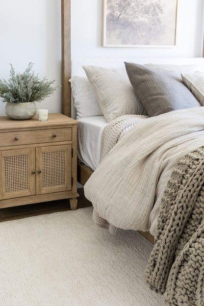 Neutral bedroom with wooden nightstand, potted plant, candle, beige bedding, chunky gray knit throw, framed tree art.