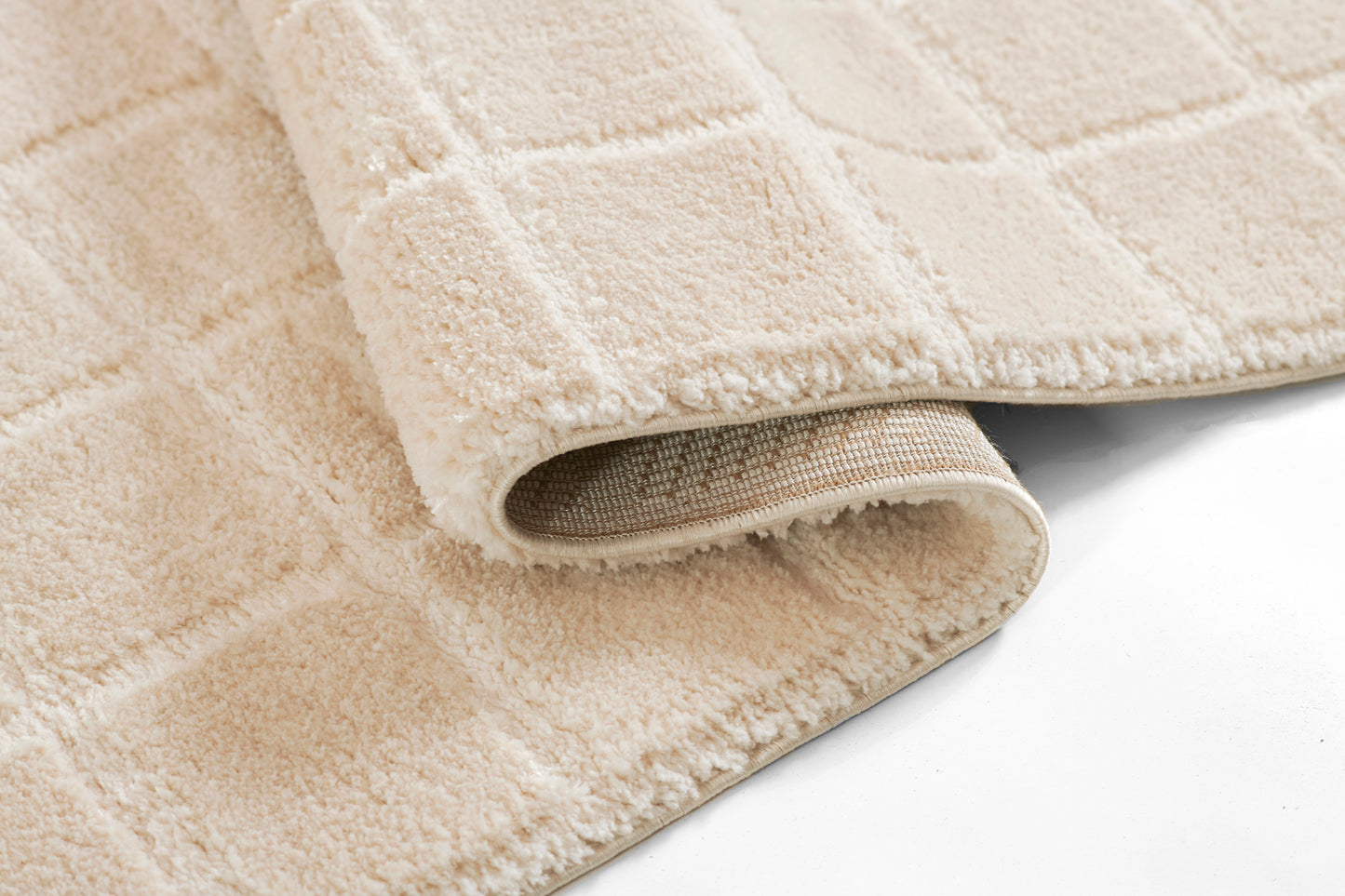 Beige plush rug with a subtle grid-pattern, soft shag pile and rolled edge revealing woven backing.