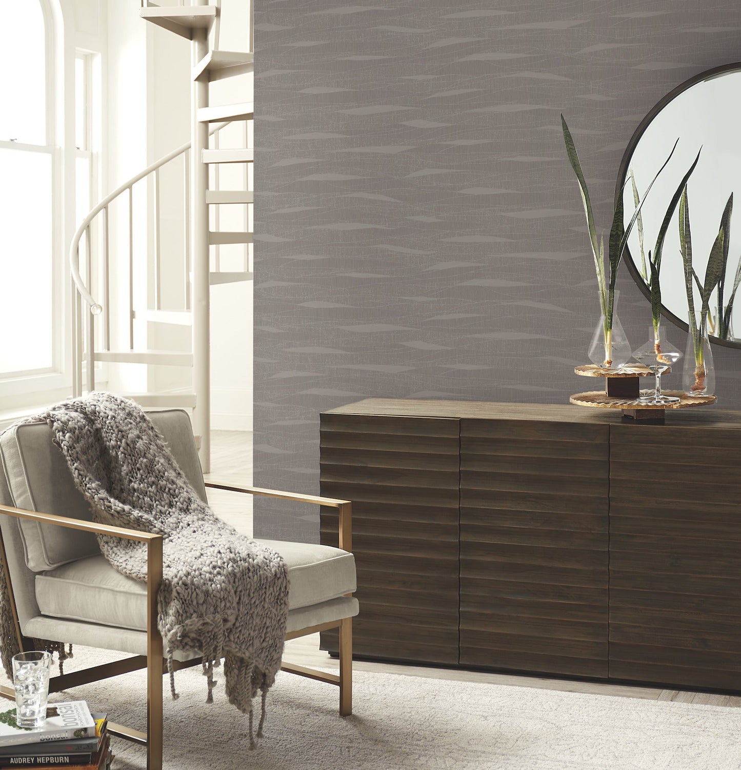 Contemporary living room with wallpapered walls featuring a subtle pattern, gray upholstered armchair with wooden frame, knit throw, dark slatted sideboard, mirror, and plant.