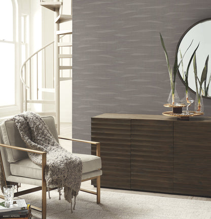 Contemporary living room with wallpapered walls featuring a subtle pattern, gray upholstered armchair with wooden frame, knit throw, dark slatted sideboard, mirror, and plant.