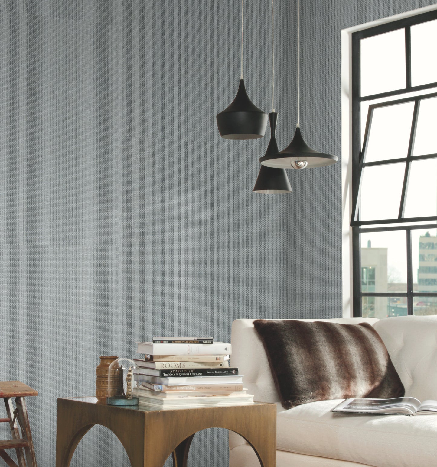 Wall covered with white Metro Tile geometric wallpaper in whites and off-whites, in a modern living room with black pendant lamps, white sofa, brown fur throw, wooden side table & books.