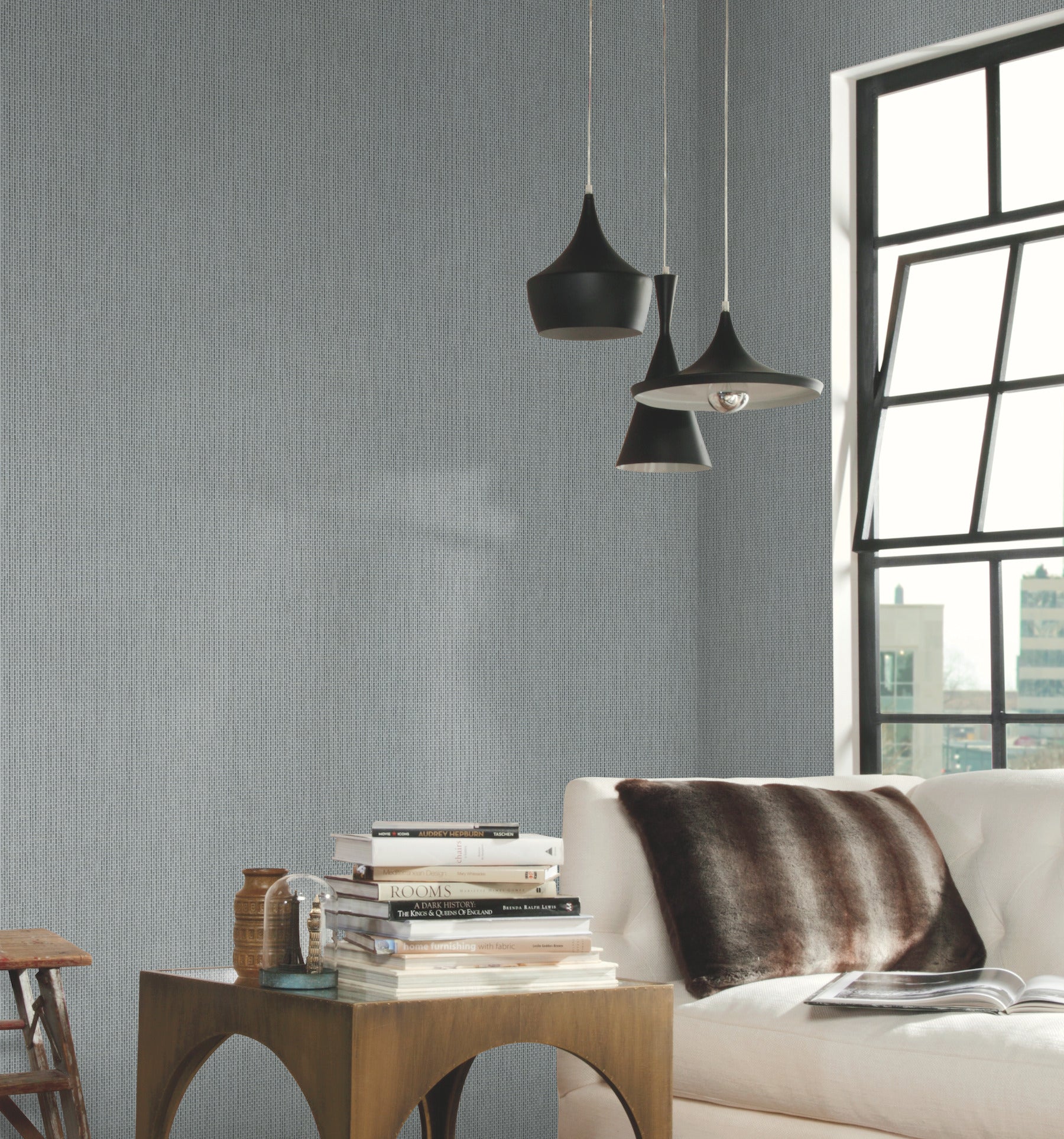 Wall covered with white Metro Tile geometric wallpaper in whites and off-whites, in a modern living room with black pendant lamps, white sofa, brown fur throw, wooden side table & books.