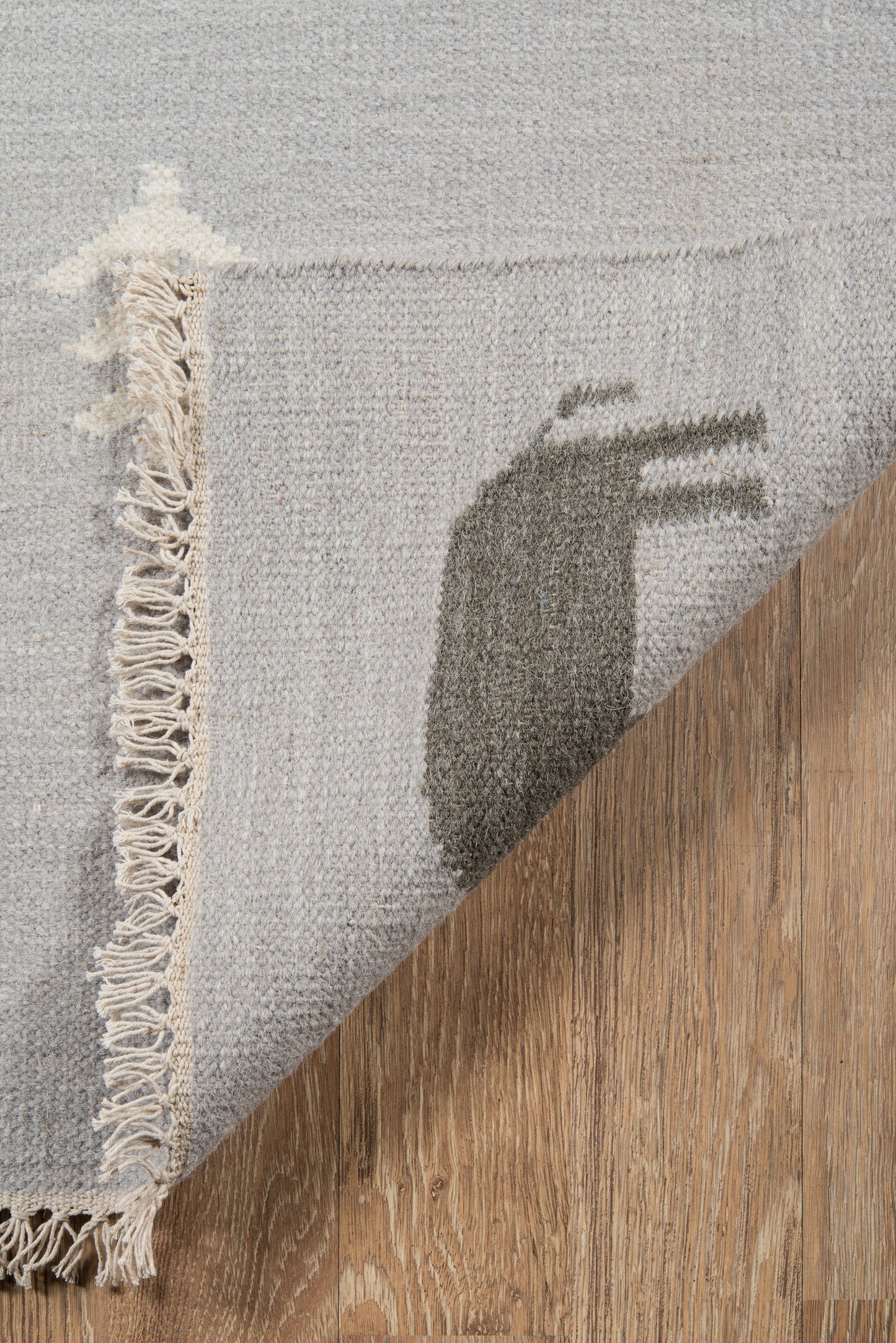 Grey woven area rug with fringed edge, folded corner reveals a dark abstract oval motif on wood floor.