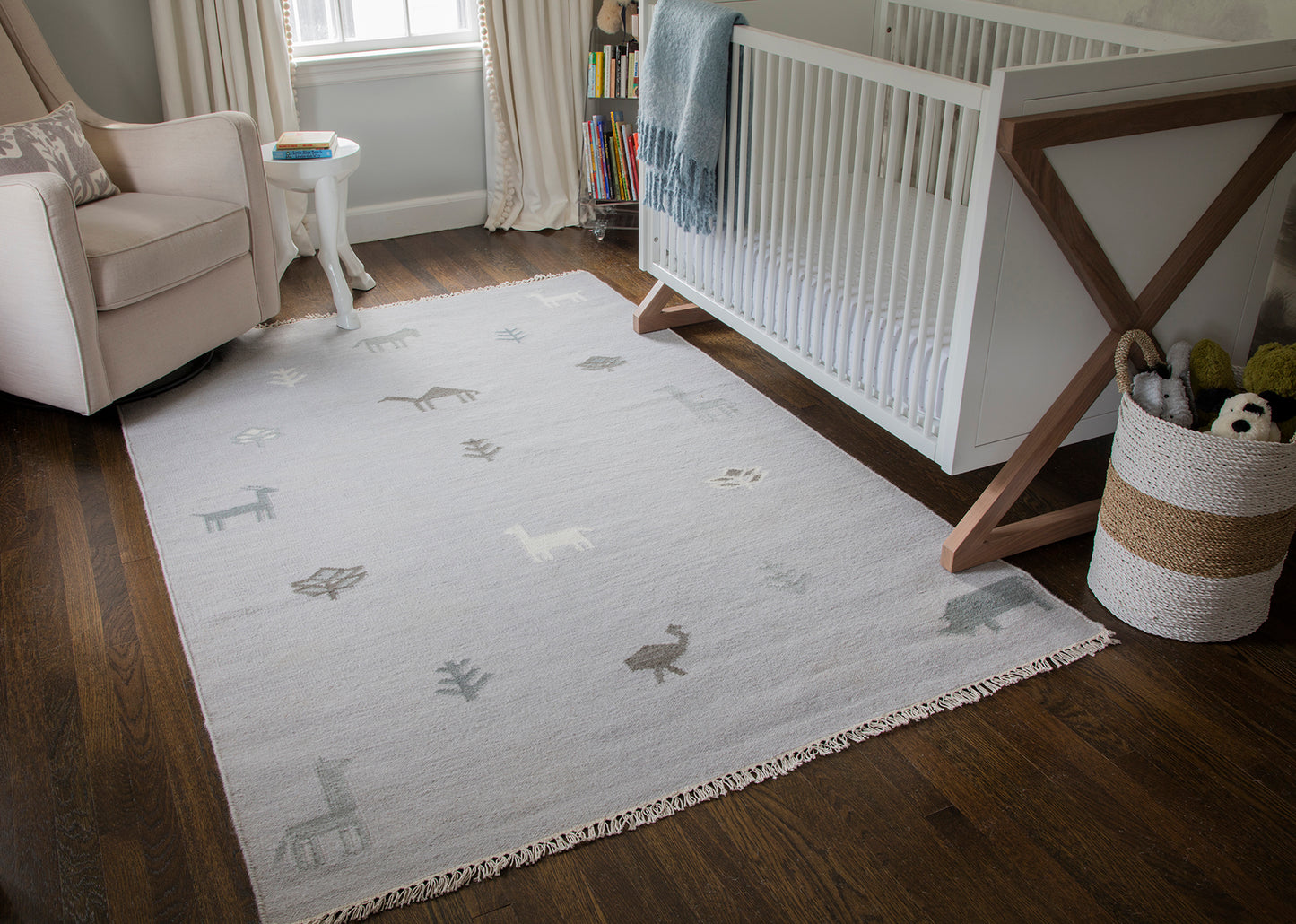 Soft light gray nursery rug with subtle animal silhouettes and fringed edges on dark wood floor.
