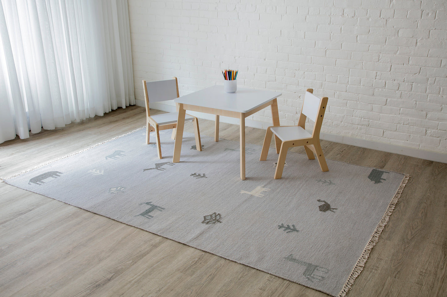 Kids' white table with natural wood legs and two matching chairs on a light gray rug with abstract shapes.
