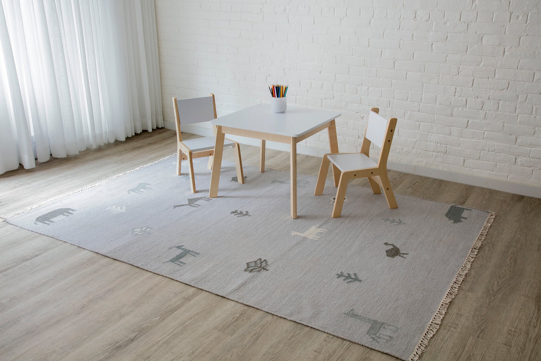 Kids' white table with natural wood legs and two matching chairs on a light gray rug with abstract shapes.