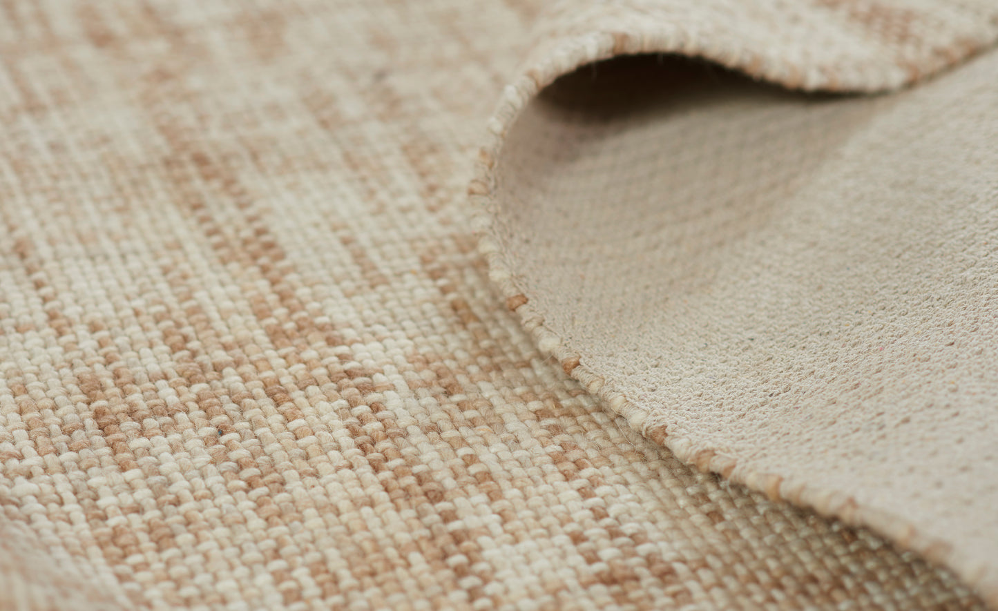 Beige woven rug with cream and tan fibers; rolled corner reveals soft suede backing for natural, cozy home decor.