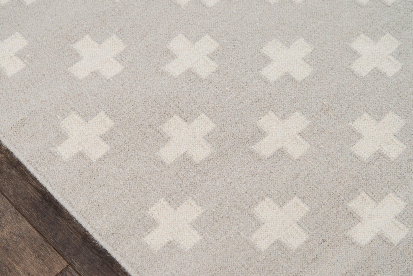 Light gray rug with white X-shaped cross pattern, edge resting on dark wood flooring.