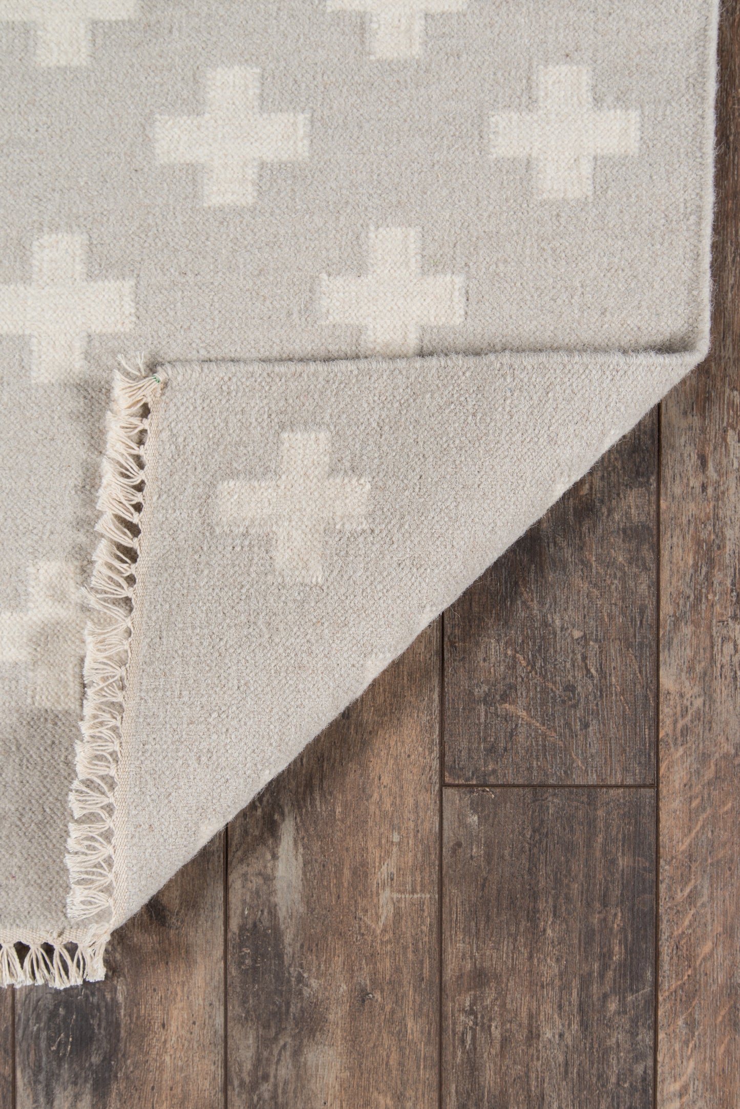 Beige-gray area rug with white plus-cross motifs, textured weave, fringed edge, folded on dark wooden floor.