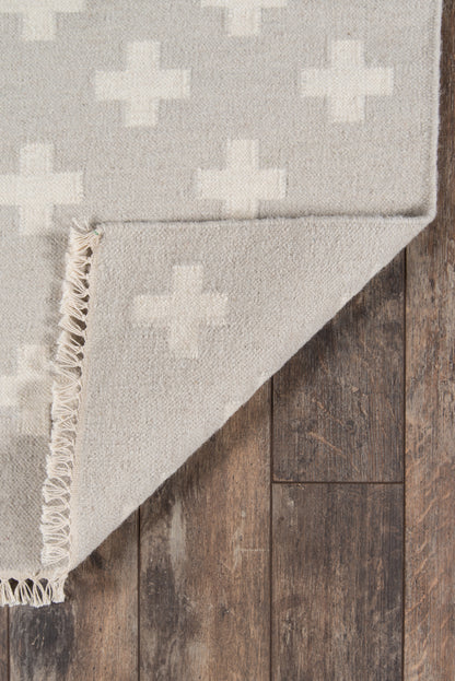 Light gray rug with white plus-pattern, fringed edge, on dark wood floor.