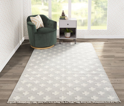 Grey area rug with white plus motifs and fringe, beside a green velvet round chair and a white side table by sunlit windows.