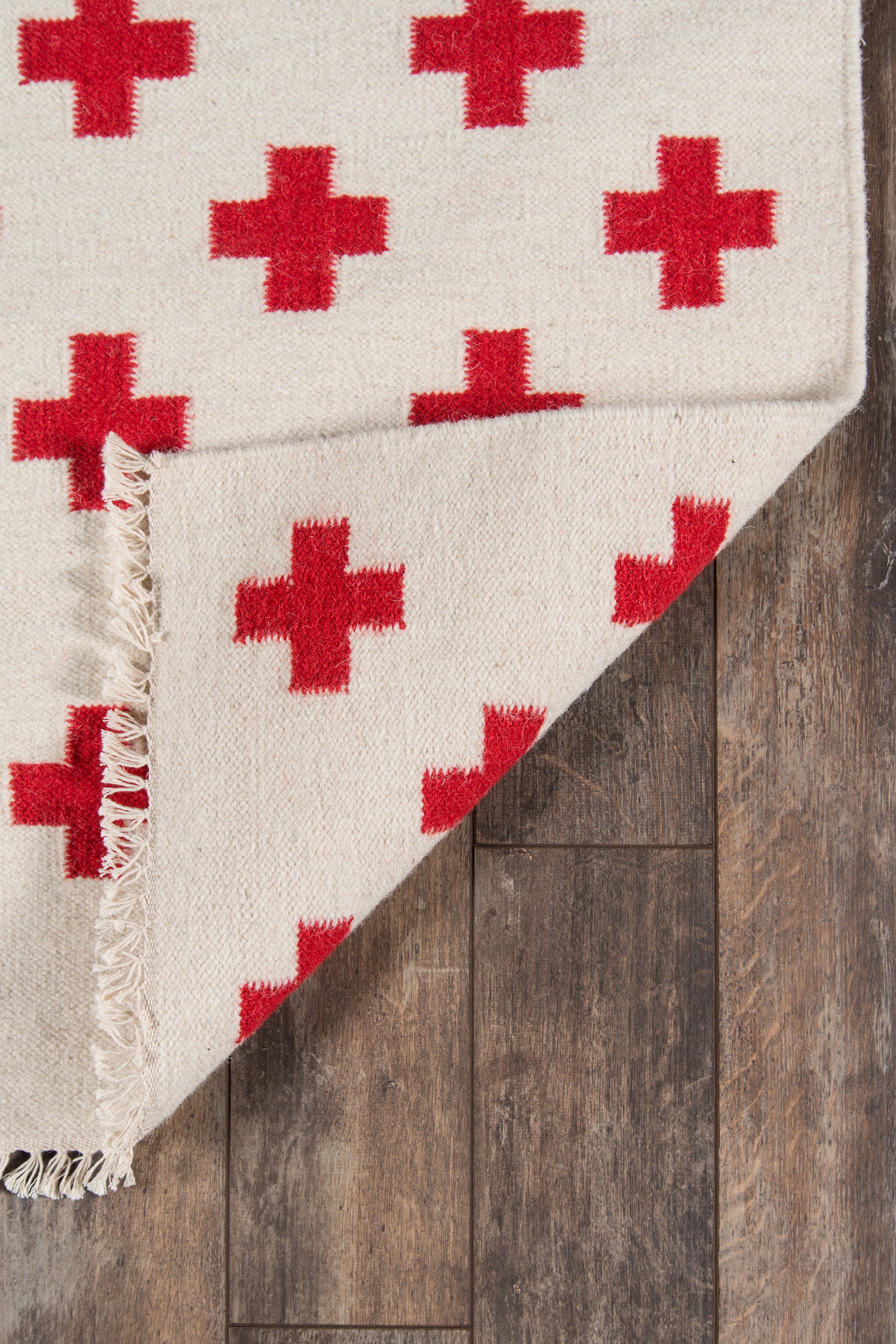 Beige woven rug with red plus pattern and fringed edges on dark wood floor.