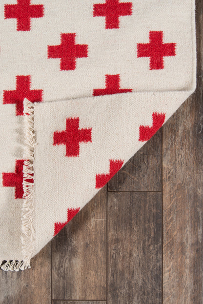 Beige woven rug with red plus motifs and fringed edge on dark wooden floor.