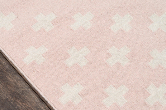 Soft pink area rug with white X-shaped motifs on a wooden floor; modern, cozy home decor.