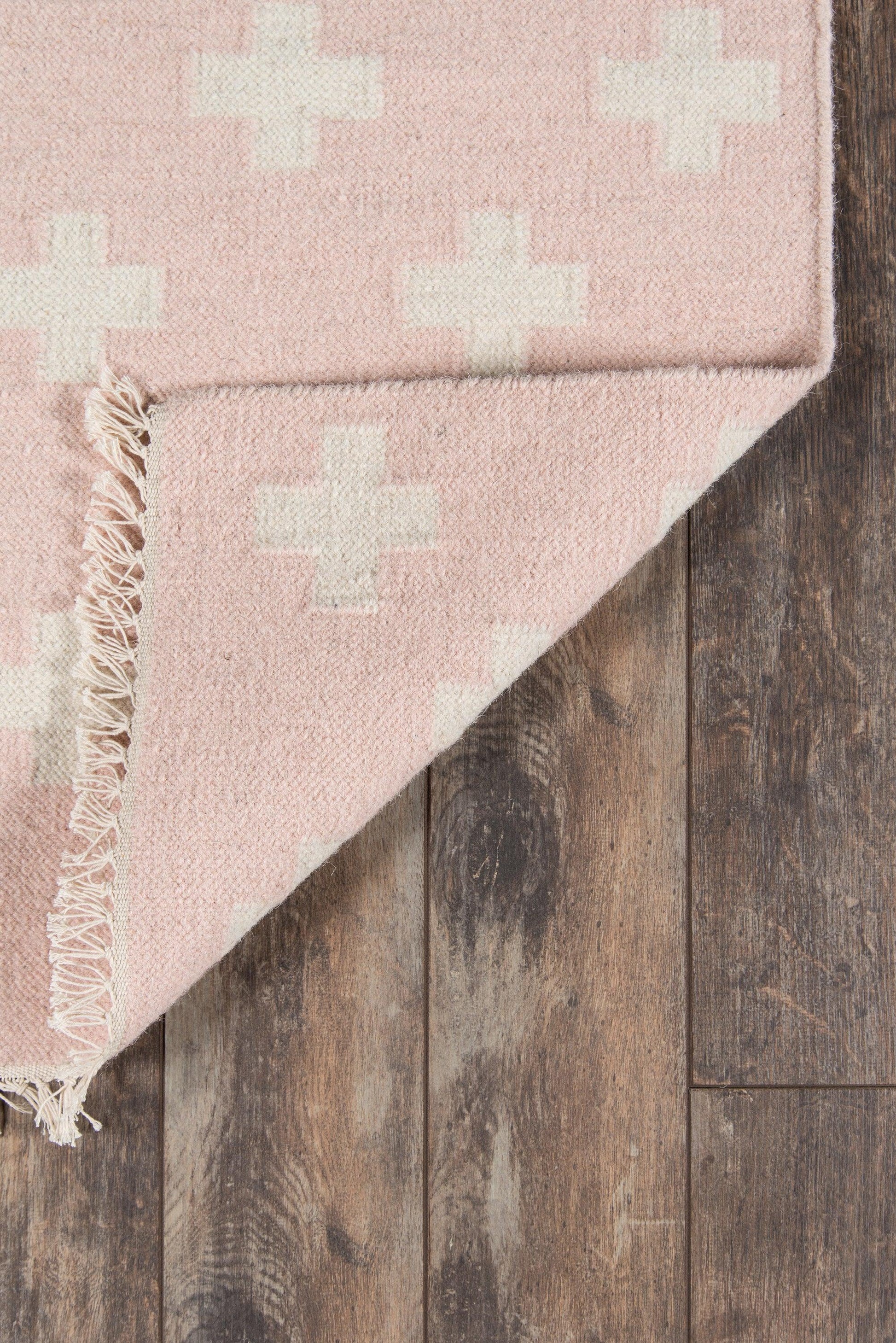 Soft pink woven rug with cream cross patterns, fringe edge, folded corner on dark wood floor.