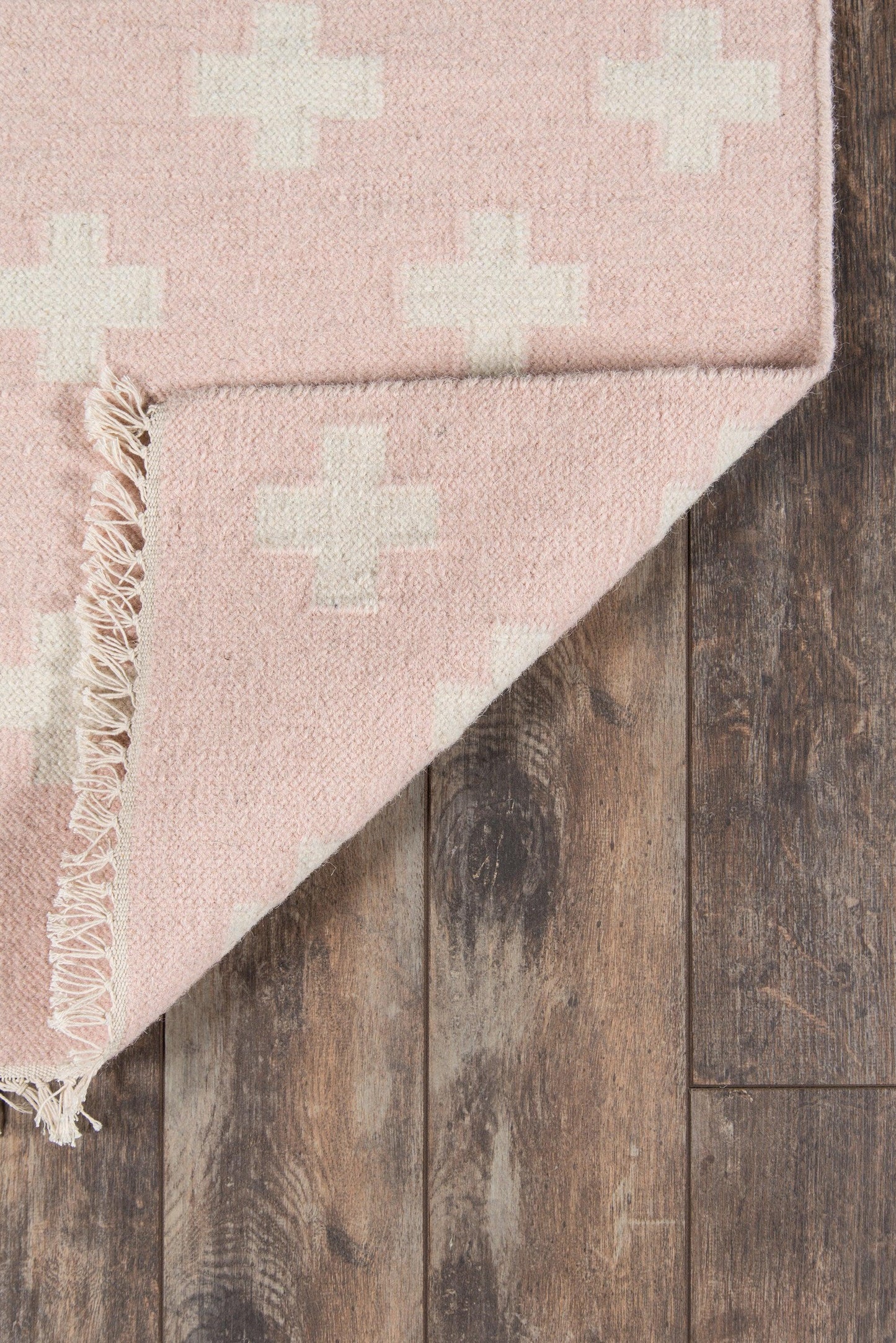 Pink rug with white cross motifs, fringed edge, on dark wood floor.