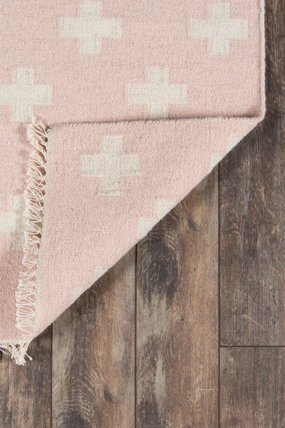 Pink rug with white cross motifs, fringed edge, on dark wood floor.