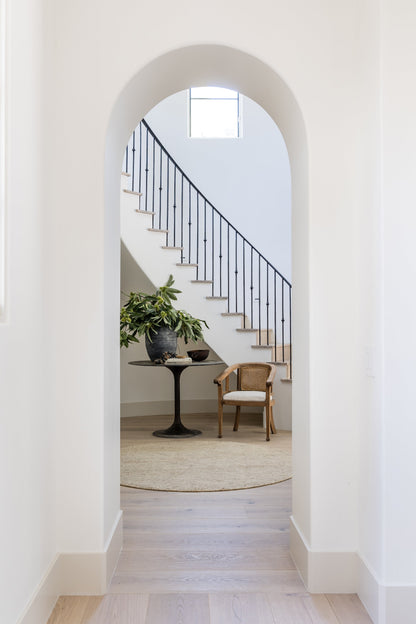 Arched doorway to a bright hallway with a staircase, black railing, round pedestal table with a plant, a rattan chair & beige rug.