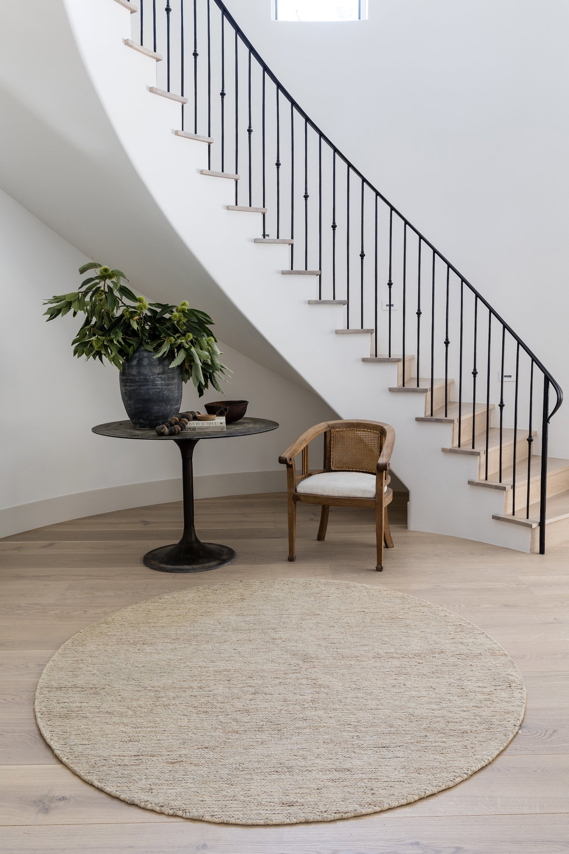 Natural beige runner rug under curved staircase with black metal railing, small wooden chair and pedestal table with a plant.