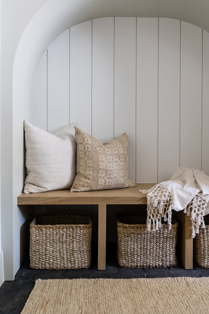 White arched alcove bench with light wood seat, beige cushions, fringed throw, and two woven baskets beneath.