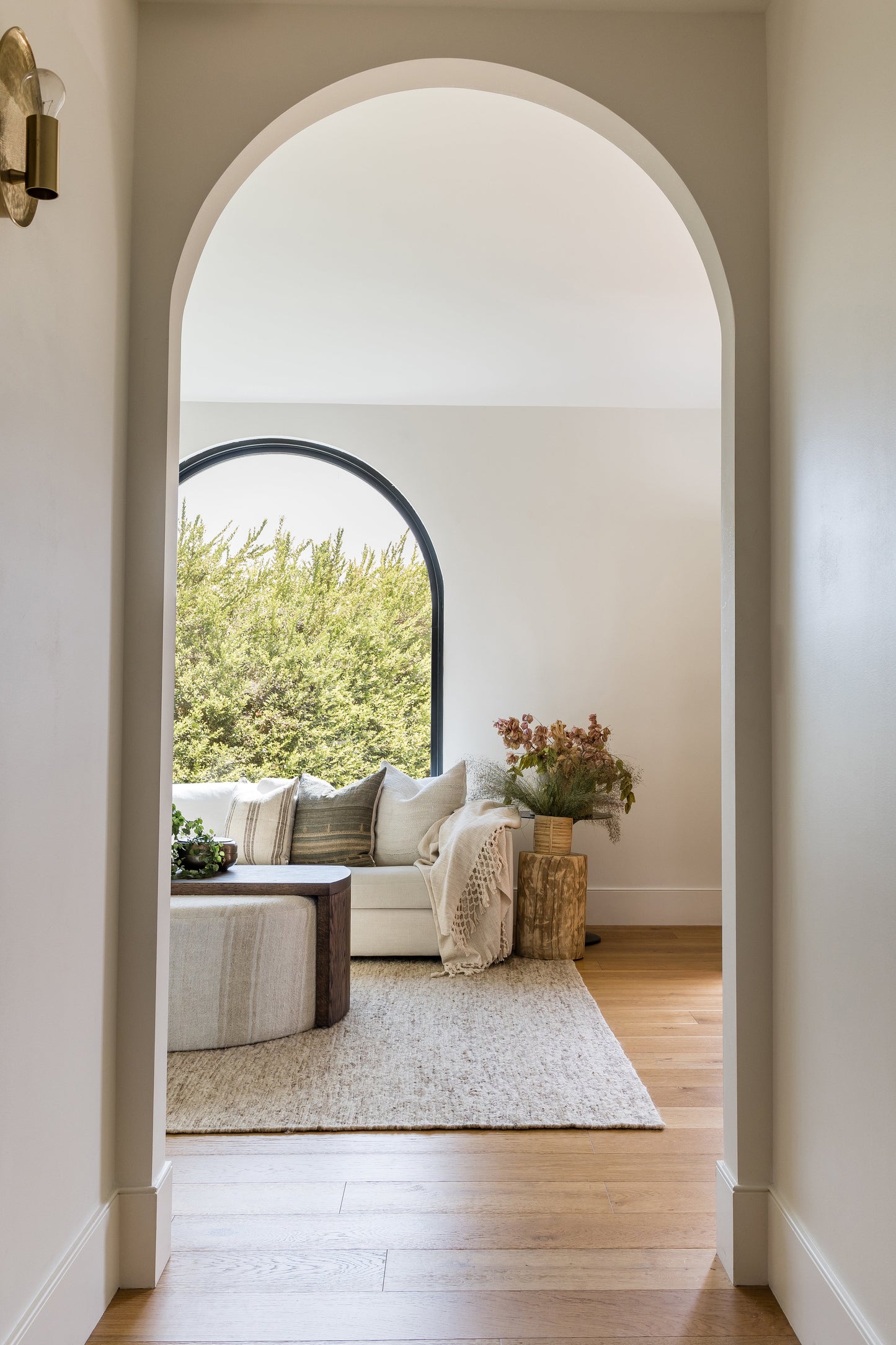 Bright living room with white walls, arched doorway and round window, beige sofa, neutral rug, wooden coffee table, plants.