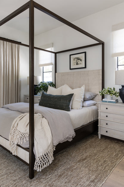 Dark wood four-poster bed with beige upholstered headboard, light gray linens, cream fringe throw, green lumbar pillow.
