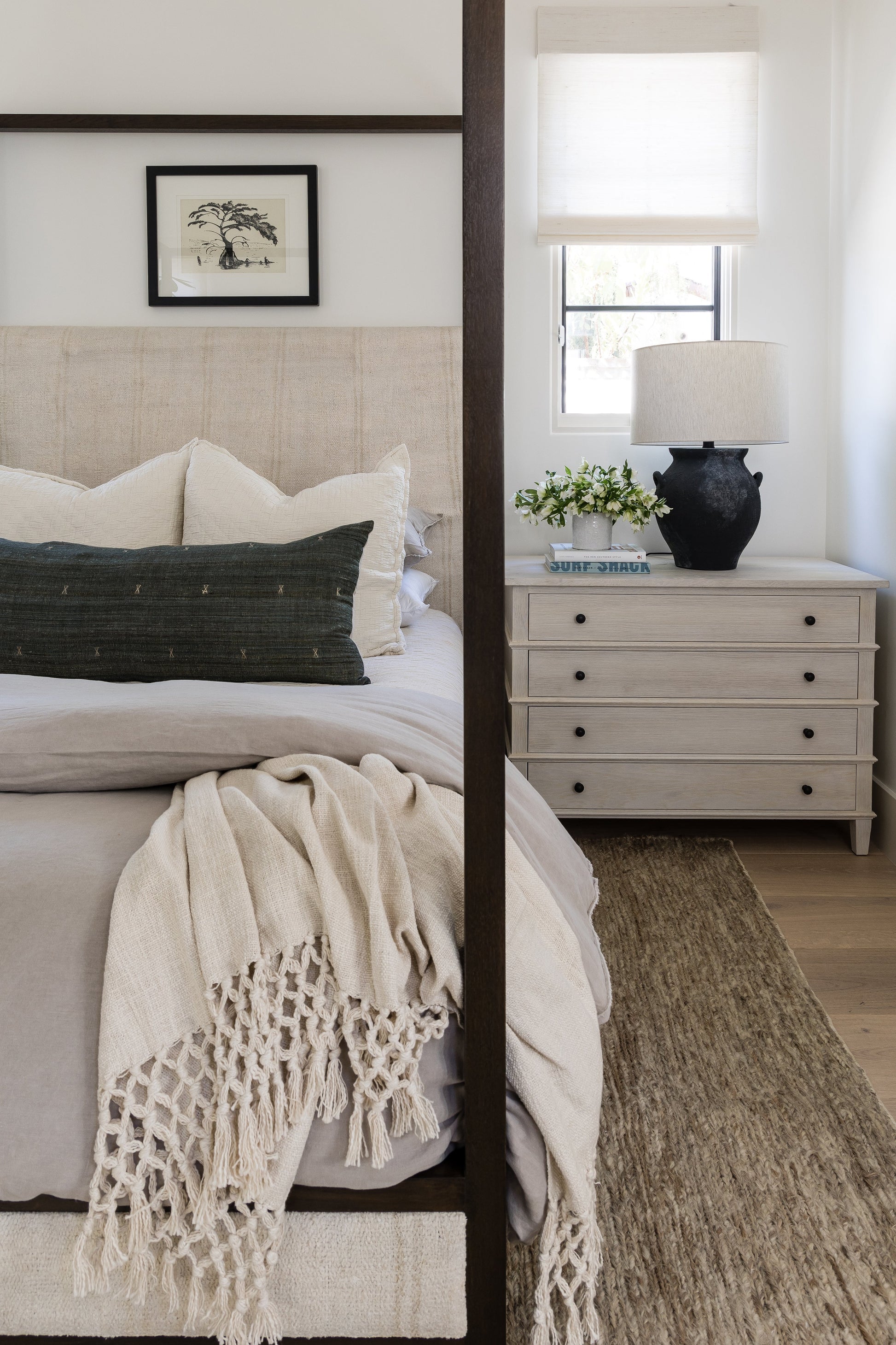 Neutral bedroom with beige upholstered bed, dark wood posts, cream bedding, green lumbar pillow, chunky knit throw, jute rug.