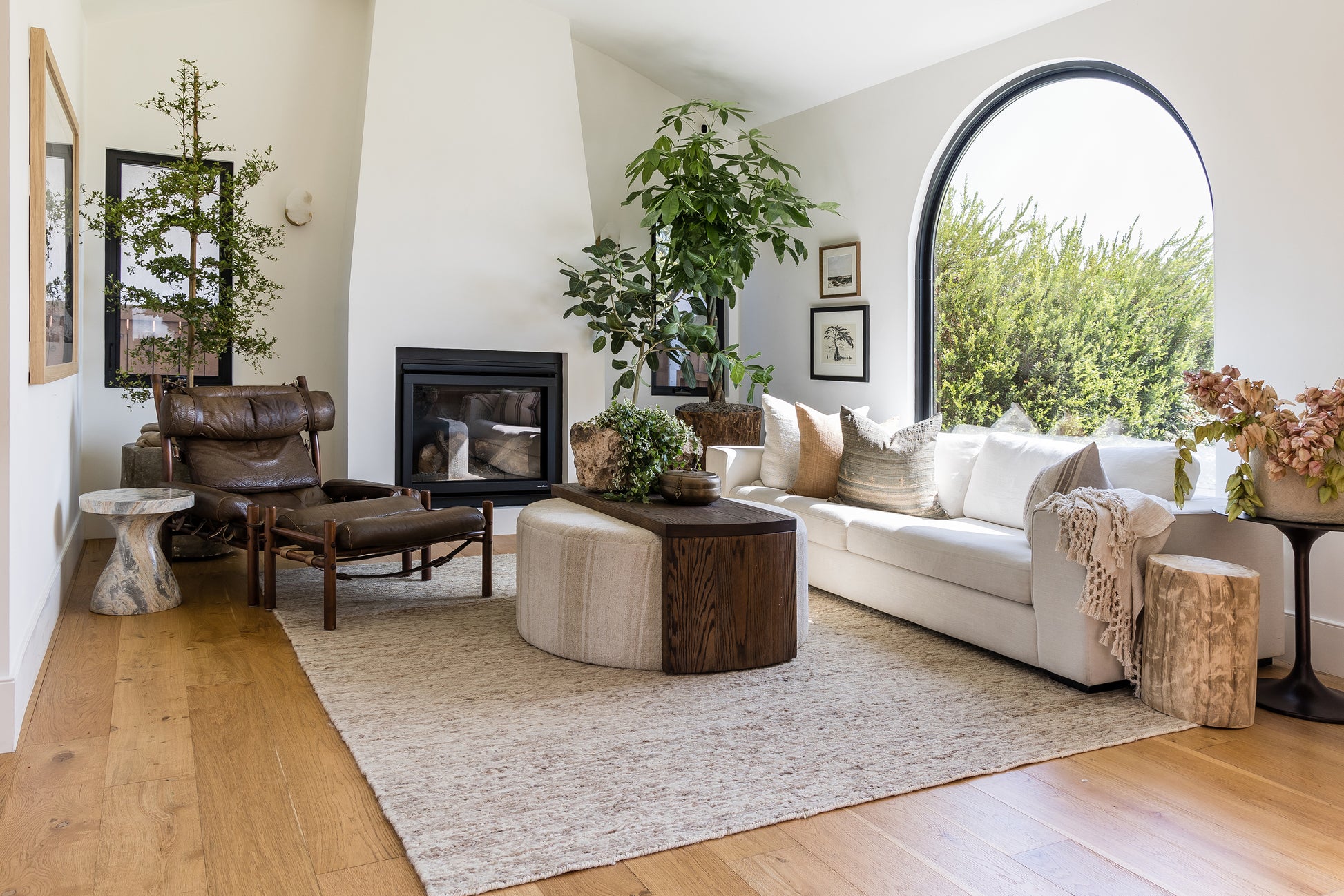 Bright modern living room with round rug, 6' X 6', white sectional, arch window, fireplace, round wooden coffee table, leather chair, greenery.