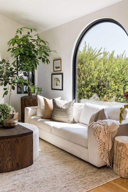 Bright living room featuring a large 10' x 14' Soumak rug, white sofa, arched window, potted plant, round wood stump tables, and neutral throw pillows.