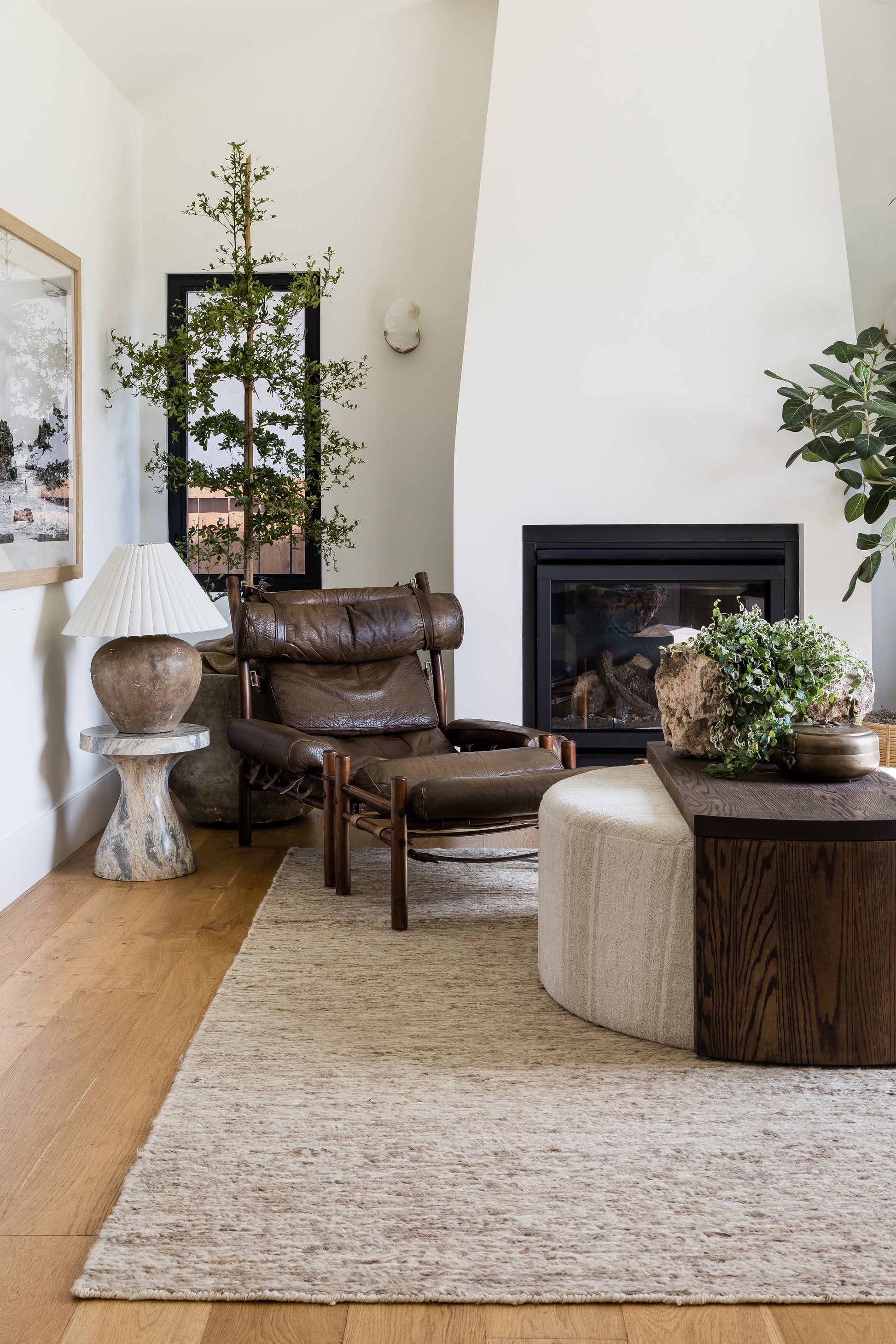 Brown leather lounge chair with ottoman, dark wood coffee table, beige textured round rug, white fireplace, and lush potted plants.