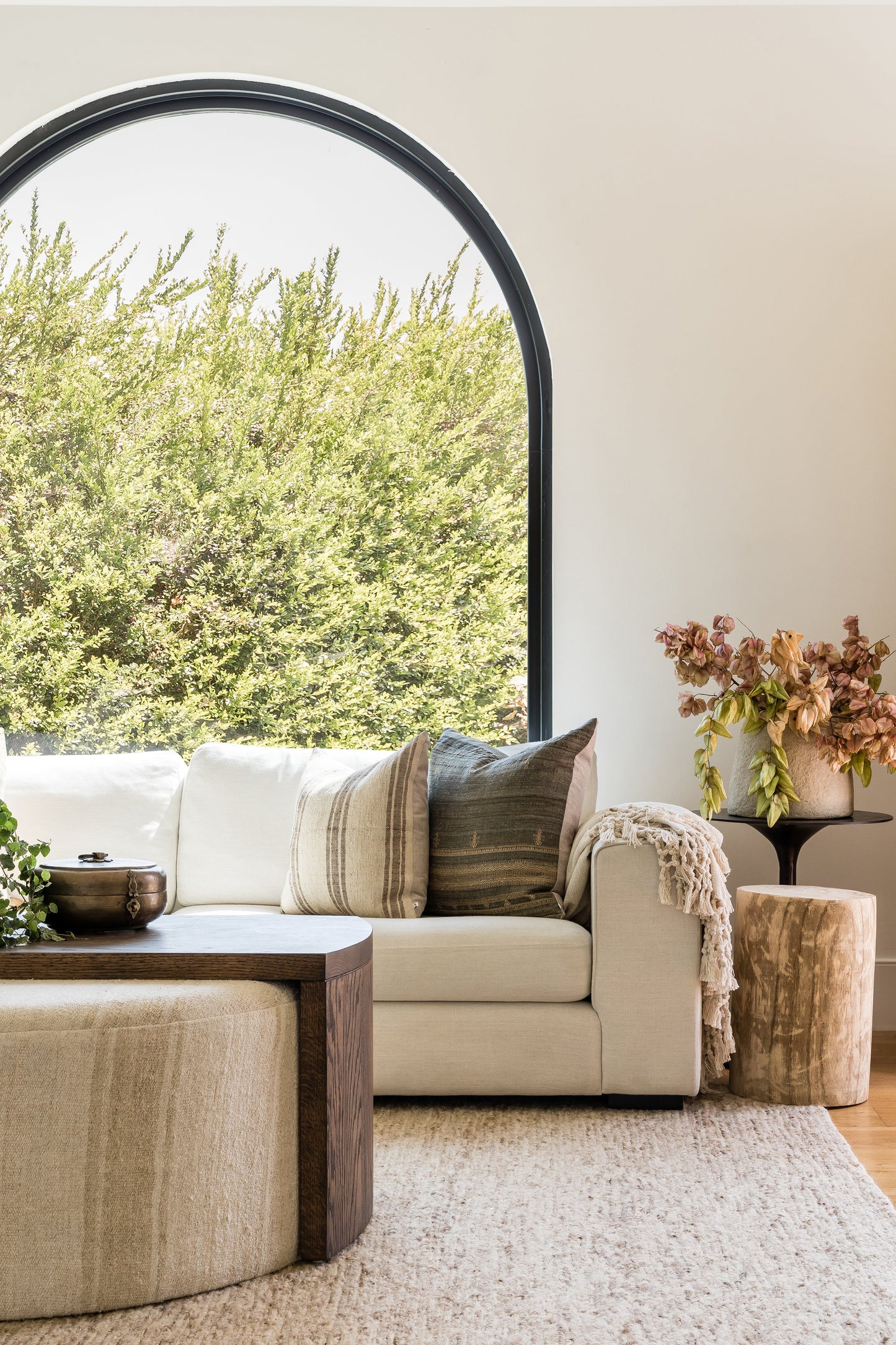 Neutral living room with a 6-foot round rug as the focal point, cream sofa with striped beige cushions, round dark wood coffee table, arched window and greenery.