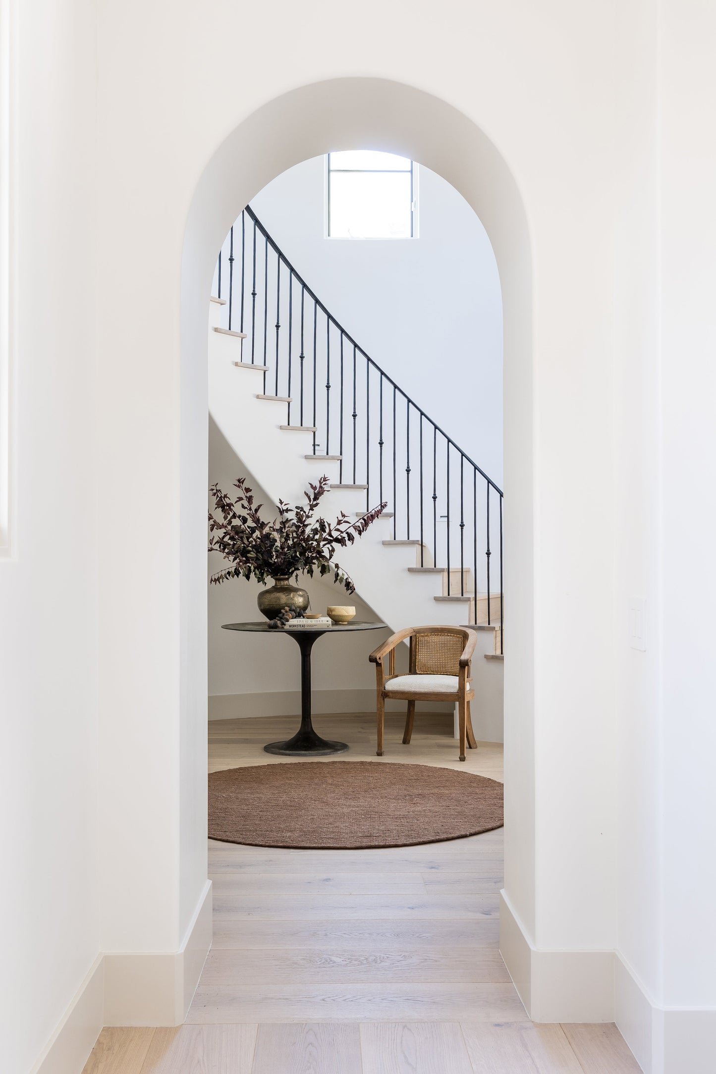Arched doorway to a bright foyer with a curved staircase, black railing, round pedestal table with flowers, wooden chair, and brown rug.