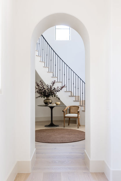 Arched doorway opens to a bright foyer with curved staircase, black iron railing, round pedestal table with a vase, wooden chair, brown rug.