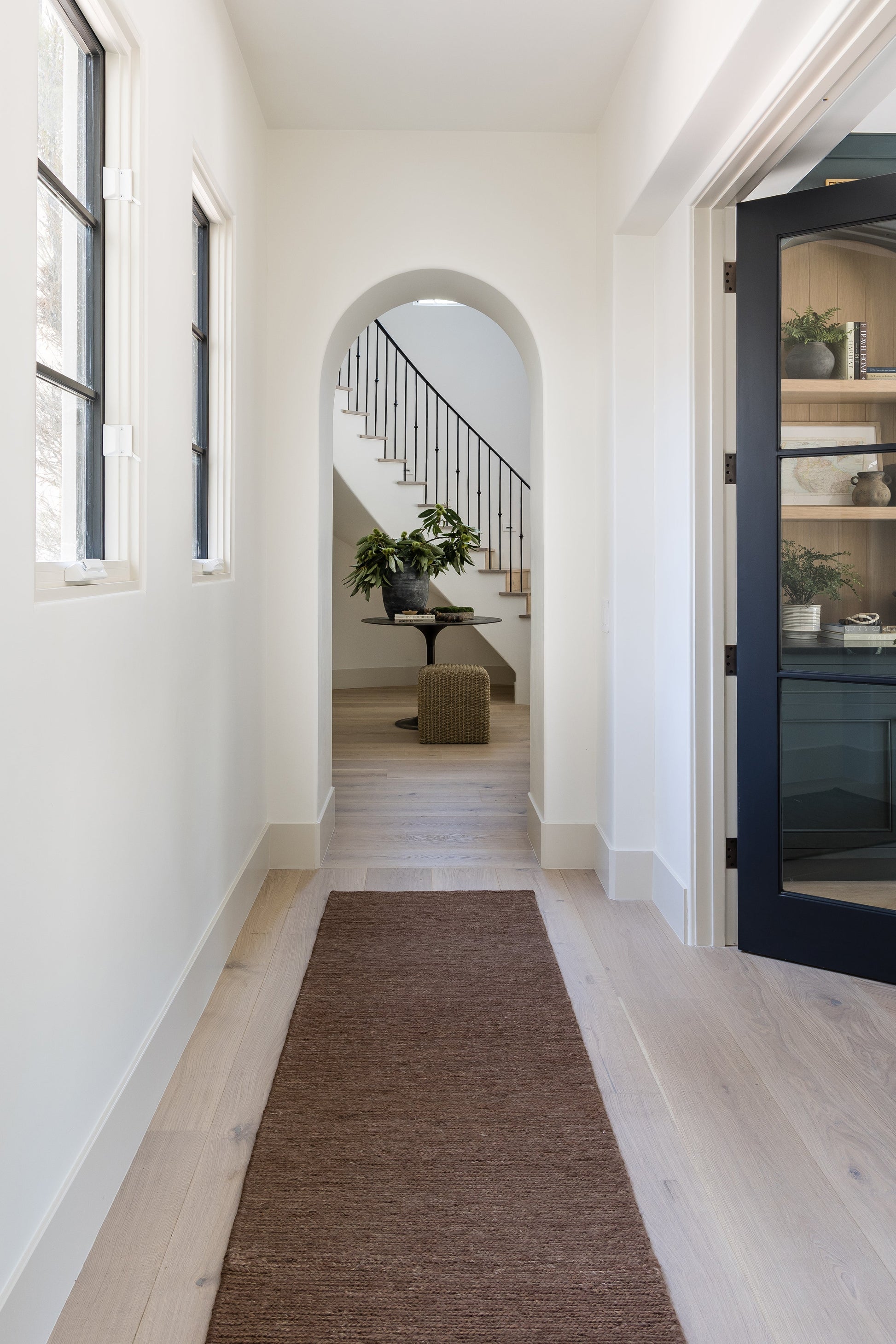 Bright hallway with light wood flooring, brown runner rug, archway to staircase with black railing, potted plant and wicker ottoman.
