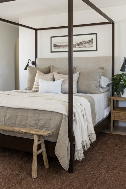 Four-poster bed with beige-gray bedding, cream throw, wooden bench at foot, matching nightstands, and framed landscape art.