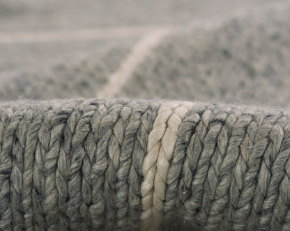 Macro view of a chunky grey wool rug edge with a cream braided stripe.