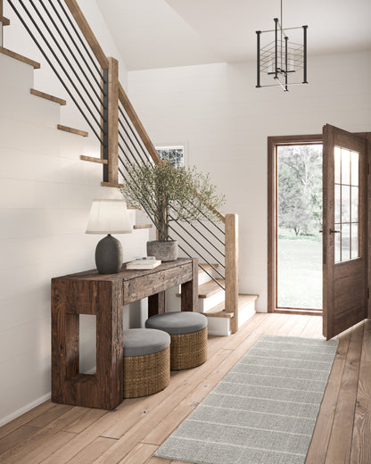 Entryway scene with a 3' x 8' rug runner on the floor, a rustic reclaimed-wood console, two gray stools with woven bases, greenery, and a black metal stair railing.