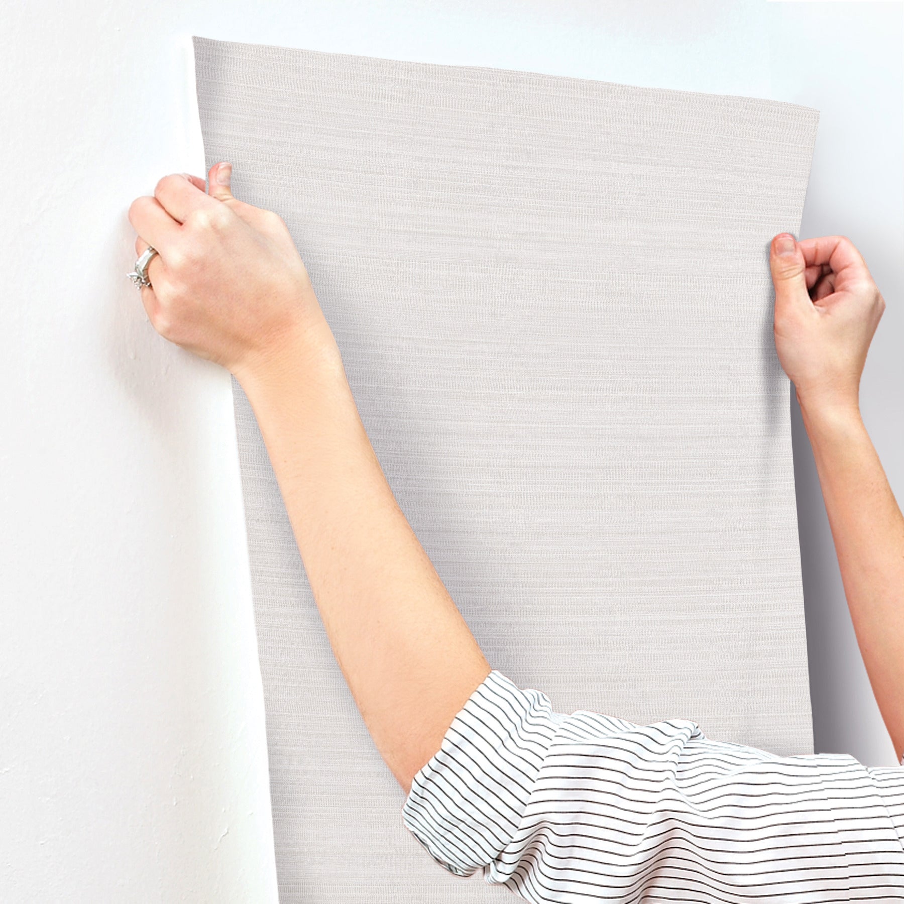 Two hands hold and smooth a light gray, textured wallpaper sheet against a wall.