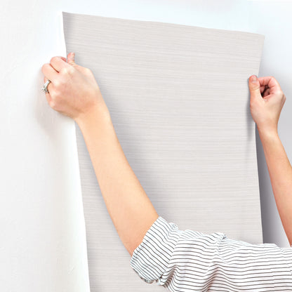 Two hands hold and smooth a light gray, textured wallpaper sheet against a wall.