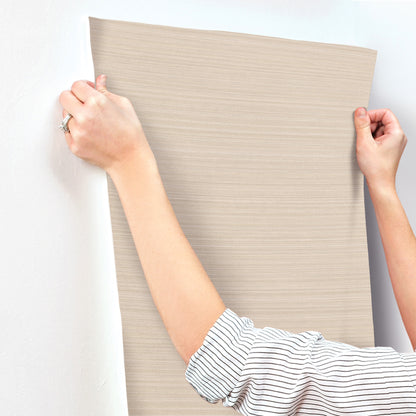 Beige textured wallpaper sheet with subtle wood-grain lines held by striped-sleeve arms, wall decor sample.